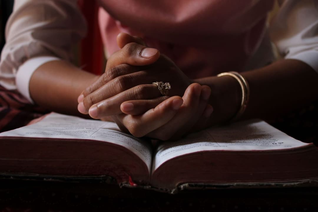 Close-up of a person's hands clasped in prayer over an open Bible.
